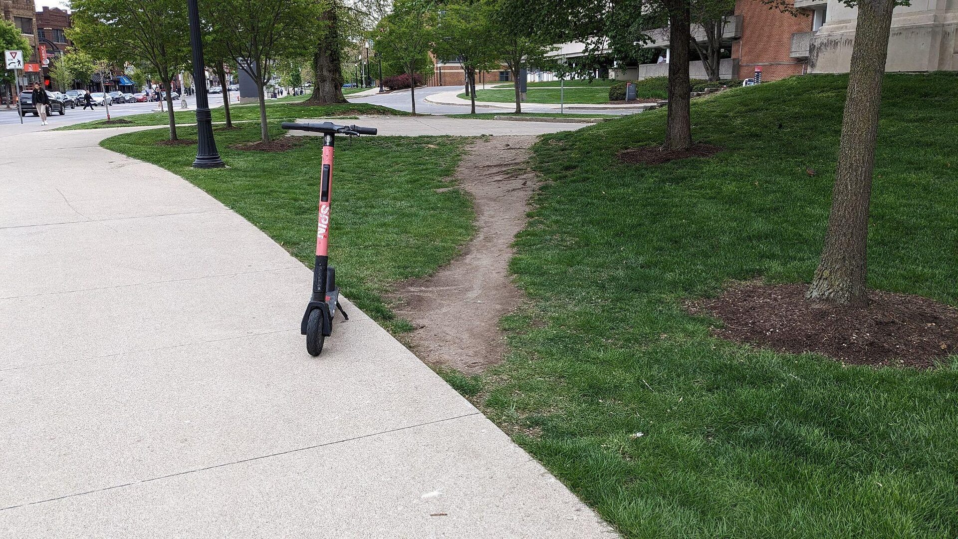 A paved campus sidewalk curves away while a worn dirt path cuts straight across the grass between two walkways. A rental scooter stands at the edge of the pavement.